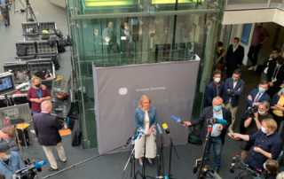 Lisa Paus steht vor einem grauen Hintergrund mit dem Bundestagslogo im Bundestag vor Mikros und Menschen