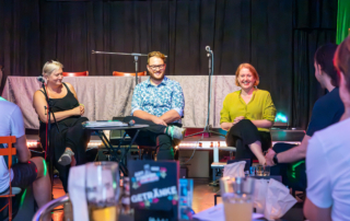 Lisa Paus sitzt zusammen mit Sebastian Weise und Maria Bormuth in einer Reihe auf Stühlen vor einer Bühne.