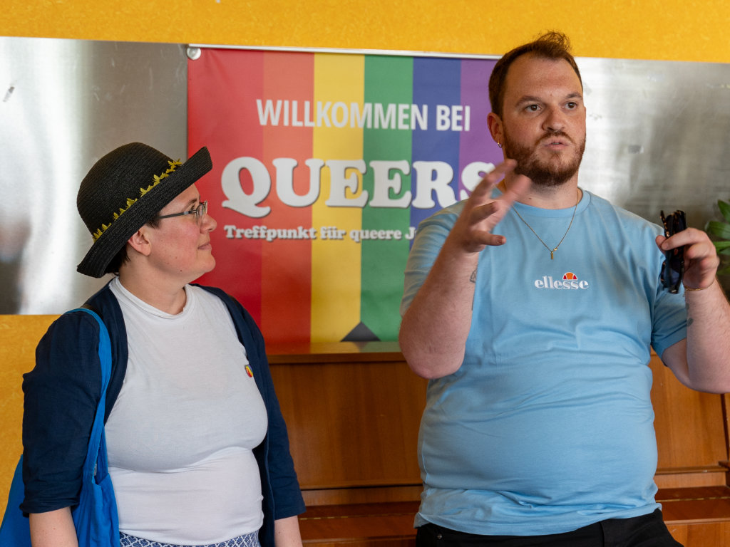Sebastian Weise und Corinna Balkow stehen vor einer Pride-Flagge.